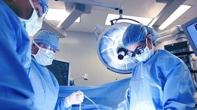 Three healthcare professionals in blue surgical gowns, masks, gloves, and caps are working in an operating room.