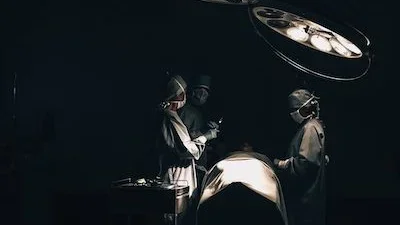 Three healthcare professionals stand next to an operating table in a mostly dark room. They are wearing blue surgical gowns, masks, gloves, and caps.