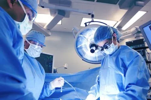 Three healthcare professionals in blue surgical gowns, masks, gloves, and caps are working in an operating room.