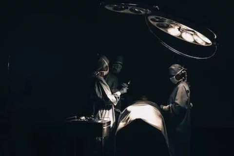 Three healthcare professionals stand next to an operating table in a mostly dark room. They are wearing blue surgical gowns, masks, gloves, and caps.