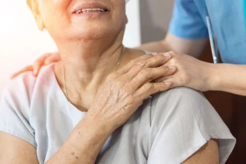 Someone is resting a hand on an older woman's shoulder. The older woman has her hand on top of the person's hand and is looking up at them.